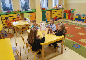 Dzieci siedzą przy stolikach, rozmawiają i bawią się podczas porannego dyżuru w sali przedszkolnej.
