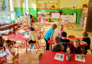 Dzieci siedzą przy stolikach i wykonują zadania w kartach pracy.