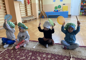 dzieci siedzą na dywanie, na głowie mają czapeczki w dłoniach trzymają przybory kuchenne