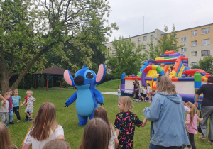 Duża maskotka Sticha otoczona przez roześmiane dzieci. Maluchy przytulają bajkowego bohatera. W tle kolorowe dekoracje i letnia, radosna atmosfera święta.