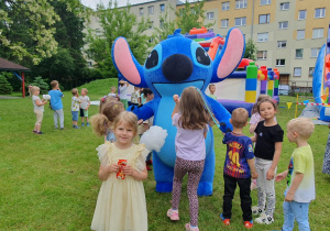 Duża maskotka Sticha otoczona przez roześmiane dzieci. Maluchy przytulają bajkowego bohatera. W tle kolorowe dekoracje i letnia, radosna atmosfera święta.