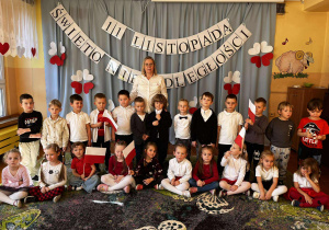 Na zdjęciu widać dzieci z grupy IV zgromadzone w sali – stoją równo, odświętnie ubrane, z biało-czerwonymi kotylionami przypiętymi do ubrań. Wspólnie pozują do zdjęcia