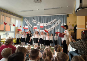 występ najstarszej grupy – dzieci prezentują inscenizację o drodze Polski do niepodległości. W tle widnieje dekoracja w barwach narodowych: biało-czerwona flaga, godło oraz napisy „11 listopada – Święto Niepodległości”.