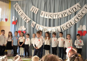 Na następnym zdjęciu widzimy dzieci w białych bluzkach i czarnych spodniach, które recytują wiersze o Polsce.