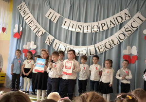 występ najstarszej grupy – dzieci prezentują inscenizację o drodze Polski do niepodległości. W tle widnieje dekoracja w barwach narodowych: biało-czerwona flaga, godło oraz napisy „11 listopada – Święto Niepodległości”.
