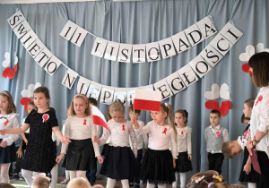 Na następnym zdjęciu widzimy dziewczynki w białych bluzkach i czarnych spódniczkach, które recytują wiersze o Polsce.