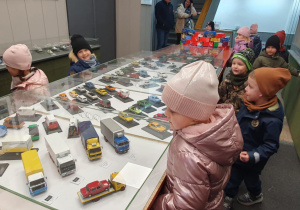 Dzieci z zaciekawieniem oglądają modele pojazdów z okresu PRL-u, ustawione na stoliku w sali.
