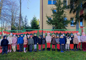 Przedszkolaki ubrane są w czerwone stroje, podkreślając jedność i zaangażowanie w kampanię „Dzieciństwo bez przemocy”. Na twarzach dzieci widać radość i dumę z udziału w akcji promującej budowanie bezpiecznych, pełnych szacunku relacji.