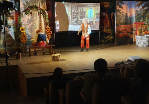 Na zdjęciu widać scenę przedstawienia teatralnego „Pinokio”. Aktor zwraca się do widowni, zadając pytanie dzieciom, a po lewej stronie na ławce siedzi tyłem postać krasnala, będąca elementem scenografii.