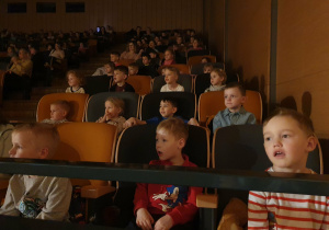 Na zdjęciu widać dzieci siedzące na krzesełkach podczas oglądania spektaklu teatralnego. Dzieci są zwrócone w stronę sceny i z uwagą obserwują przedstawienie.