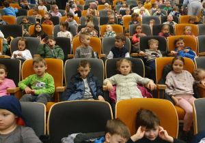 Na zdjęciu widać dzieci siedzące na krzesełkach na widowni przed rozpoczęciem spektaklu. Dzieci rozmawiają ze sobą i oczekują na rozpoczęcie przedstawienia.
