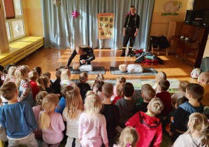 Pokaz resuscytacji na fantomie.Na zdjęciu pan ratownik przeprowadza resuscytację na fantomie, pochylając się nad nim, a drugi ratownik stoi z tyłu, obserwuje. Dzieci siedzą na dywanie i uważnie przyglądają się całej sytuacji. Scena pokazuje edukacyjne ćwiczenia z zakresu pierwszej pomocy w bezpiecznej i pouczającej atmosferze.