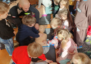 Dziecko ćwiczy resuscytację na fantomie pod okiem instruktora, pozostałe dzieci obserwują.