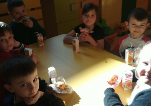 Na zdjęciu dzieci siedzą przy stoliku w przedszkolu. Jedzą słodkie babeczki i piją soki. Dzieci są uśmiechnięte i skupione na swoim poczęstunku. Atmosfera jest radosna i przyjazna.