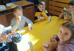 Na zdjęciu dzieci siedzą przy stoliku w przedszkolu. Każde dziecko ma przed sobą babeczkę i kubek soku. Dzieci uśmiechają się i rozmawiają ze sobą podczas jedzenia.