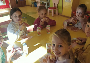 Dzieci siedzą przy stoliku i jedzą słodkie babeczki. Są wesołe i skupione na poczęstunku, a atmosfera jest przyjazna i radosna.