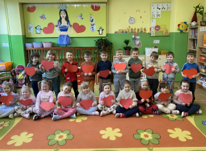 Na zdjęciu dzieci siedzą na dywanie, każde trzyma w dłoniach czerwone serce. Za nimi widać wiosenną dekoracje w kolorach zielonych. Dzieci patrzą w stronę aparatu, uśmiechają się i tworzą ra