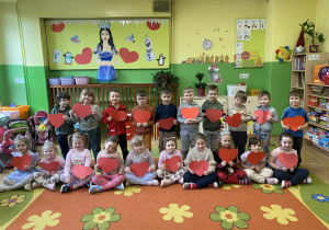 Na zdjęciu dzieci siedzą na dywanie, każde trzyma w dłoniach czerwone serce. Za nimi widać wiosenną w kolorach zielonych. Dzieci patrzą w stronę aparatu, uśmiechają się i tworzą radosną, kolorową scenę.