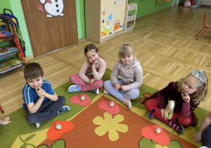 Na zdjęciu dzieci siedzą na dywanie, przed nimi leżą czerwone serca, na których poukładane są kolorowe czekoladowe jajka. Dzieci patrzą na swoje słodkości i uśmiechają się.