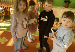 Na zdjęciu chłopcy wręczają dziewczynkom kwiatki, życząc im „100 lat”. Dziewczynki uśmiechają się, a dzieci stoją razem w grupie, tworząc radosną i pełną ciepła scenę. Widać, że moment jest pełen uśmiechów i przyjaznej atmosfery.