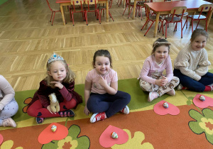 Na zdjęciu widać cztery dziewczynki siedzące na dywanie. Uśmiechają się w stronę aparatu. Przed nimi leżą czerwone serca. Cała scena jest radosna i pełna kolorów, pokazując wesołą atmosferę w przedszkolu.