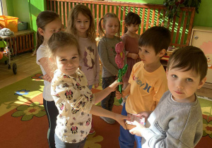 Na zdjęciu dzieci stoją w grupie. Chłopcy wręczają dziewczynkom upominki. Dziewczynki uśmiechają się, a cała scena pokazuje radosną i przyjazną atmosferę w przedszkolu.