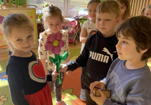 Na zdjęciu dziewczynka ubrana w czerwono-czarną sukienkę stoi w grupie dzieci. Chłopcy wręczają jej upominki – kwiatka i mały prezent. Dziewczynka uśmiecha się, a scena jest radosna i pełna ciepłej, przyjaznej atmosfery.