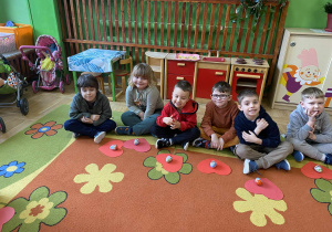 Na zdjęciu chłopcy siedzą na dywanie i uśmiechają się w stronę aparatu. Przed nimi leżą przygotowane upominki dla dziewczyn.