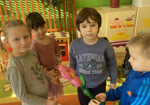 Na zdjęciu widać dwie dziewczynki i dwóch chłopców. Z przodu chłopiec w niebieskim sweterku wręcza dziewczynce różowego kwiatka.