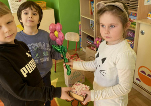 Na zdjęciu dziewczynka w białej bluzce otrzymuje od dwóch chłopców prezenty. Dziewczynka uśmiecha się, a chłopcy patrzą na nią z radością. Cała scena jest pełna uśmiechów i przyjaznej, radosnej atmosfery w przedszkolu.