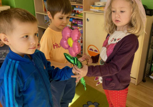 Na zdjęciu dziewczynka otrzymuje od dwóch chłopców kwiatka. Dziewczynka uśmiecha się, a chłopcy patrzą na nią z radością. Scena jest pełna ciepłej, przyjaznej i radosnej atmosfery.