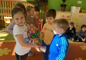 Dziewczynka otrzymuje mały prezent od jednego z chłopców. Trzyma go w rękach z zaciekawieniem i uśmiechem. W tle widać innych uczniów obserwujących moment. Zdjęcie 3: