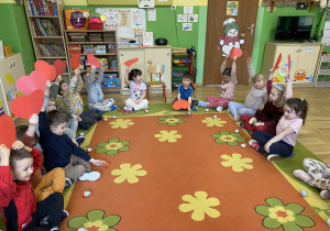 Dzieci siedzą na dywanie w siadzie po turecku. Przed nimi leżą upominki przygotowane dla dzieci. Patrzą w stronę aparatu i uśmiechają się. Scena jest radosna i pełna przyjaznej atmosfery.