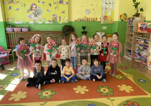 Na zdjęciu cała grupa dzieci pozuje do zdjęcia. Z tyłu stoją dziewczynki, trzymając w dłoniach kolorowe kwiatki. Przed nimi, na dywanie, siedzą chłopcy w siadzie po turecku. Dzieci uśmiechają się, a scena jest radosna i pełna przyjaznej atmosfery.