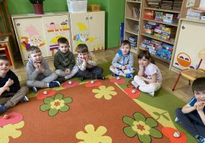 Na zdjęciu dzieci siedzą na dywanie w siadzie po turecku. Za nimi widać półkę, na której poukładane są puzzle i pomoce dydaktyczne. Dzieci patrzą w stronę aparatu, a scena pokazuje spokojną, edukacyjną i przyjazną atmosferę w przedszkolu.