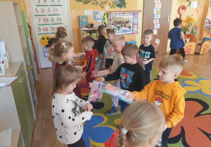 Na zdjęciu dzieci uczestniczą w obchodach Dnia Kobiet zorganizowanych przez rodziców w przedszkolu. Dziewczynki otrzymują od chłopców drobne upominki. Dzieci uśmiechają się, a scena jest pełna radości i przyjaznej atmosfery.