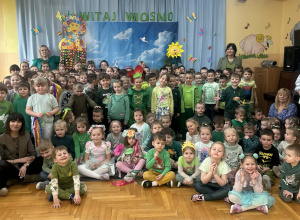 Na zdjęciu widać dzieci siedzące na podłodze w sali przedszkolnej, ubrane w zielone stroje. Za dziećmi znajduje się dekoracja z napisem „Witaj wiosno” oraz wiosenne ozdoby. Na zdjęciu widocz