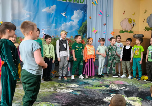 Na zdjęciu widać dzieci ubrane w wiosenne stroje, które występują przed grupą przedszkolaków. Dzieci recytują wiersze i śpiewają piosenki. W tle znajduje się wiosenna dekoracja, a po prawej stronie na ścianie widoczne są kolorowe ozdoby.