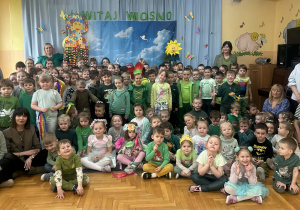 Na zdjęciu widać dzieci siedzące na podłodze w sali przedszkolnej, ubrane w zielone stroje. Za dziećmi znajduje się dekoracja z napisem „Witaj wiosno” oraz wiosenne ozdoby. Na zdjęciu widoczna jest cała społeczność przedszkolna podczas wspólnego świętowania powitania wiosny.