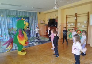 Na zdjęciu widać dzieci uczestniczące w zajęciach ruchowo-tanecznych w sali przedszkolnej. Przedszkolaki stoją w półkolu i wykonują proste ćwiczenia, podskakując oraz unosząc ręce. Przed nimi znajduje się duża, kolorowa maskotka w kształcie papugi, która prowadzi zabawę i zachęca dzieci do aktywności. W tle widoczna jest dekoracja z napisem „Witaj Wiosno” oraz plansza przedstawiająca wiosenny krajobraz. Ściany sali ozdobione są elementami tematycznymi, takimi jak nutki, zwierzątka i kwiaty. Po prawej stronie znajdują się drabinki gimnastyczne z przypiętymi papierowymi kwiatami, a także kolorowe pomoce dydaktyczne. Całość ukazuje radosną atmosferę wspólnej zabawy, ruchu i integracji dzieci podczas zajęć.