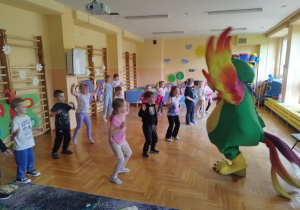 Na zdjęciu widoczna jest grupa dzieci biorących udział w zajęciach ruchowych w sali przedszkolnej. Dzieci stoją w rozproszeniu na drewnianej podłodze i wykonują proste ćwiczenia ruchowe, unosząc ręce i poruszając się w rytmie zabawy. Przed nimi znajduje się duża, kolorowa maskotka w kształcie papugi, która prowadzi aktywność i zachęca dzieci do wspólnego tańca. Sala jest przestronna i dobrze oświetlona, z jasnymi ścianami ozdobionymi elementami dekoracyjnymi, takimi jak słońce, chmurki i kolorowe kształty. Po bokach widoczne są drabinki gimnastyczne oraz sprzęty i materiały wykorzystywane podczas zajęć. Zdjęcie przedstawia radosną atmosferę wspólnej zabawy, aktywności fizycznej i integracji dzieci.