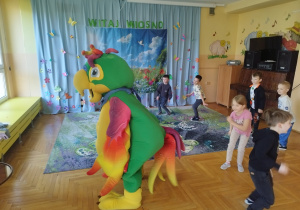 Na zdjęciu dzieci biorą udział w zajęciach ruchowych w sali przedszkolnej. Pod kierunkiem kolorowej maskotki papugi wykonują proste ćwiczenia i podskoki. W tle widoczna jest wiosenna dekoracja z napisem „Witaj Wiosno”.