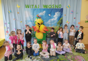 Na zdjęciu widoczna jest grupa dzieci z przedszkola pozujących do wspólnego zdjęcia wraz z dużą, kolorową maskotką w kształcie papugi. Dzieci stoją i siedzą na dywanie w sali przedszkolnej, uśmiechają się i przyjmują wesołe pozy, opierając dłonie pod brodą. W tle znajduje się dekoracja o tematyce wiosennej – jasna kurtyna ozdobiona kolorowymi motylami oraz napis „Witaj Wiosno”. Centralnie umieszczona jest plansza przedstawiająca wiosenny krajobraz z kwiatami, zielenią i niebem. Całość tworzy radosną, kolorową scenę, podkreślającą wspólną zabawę dzieci podczas zajęć.
