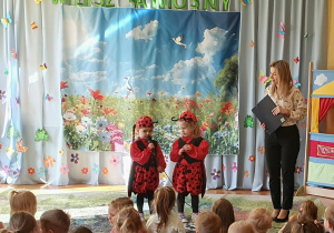Na zdjęciu widać dwie dziewczynki ubrane w stroje biedronek, które stoją na scenie i recytują wiersz. Dzieci z zaangażowaniem prezentują swój występ przed publicznością, a w tle widoczna jest kolorowa, wiosenna dekoracja.
