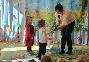 Na zdjęciu widoczny jest uroczysty moment wręczania dyplomów dzieciom, które brały udział w konkursie recytatorskim. Pani dyrektor z uśmiechem podchodzi do kolejnych uczestników i wręcza im pamiątkowe dyplomy, doceniając ich odwagę oraz piękne występy. Dzieci stoją obok pani dyrektor, z widoczną radością i dumą odbierają nagrody.