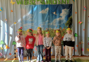 Na zdjęciu widać dzieci stojące na scenie i recytujące wiersz podczas konkursu „Wiersz dla wiosny”. W tle znajduje się kolorowa, wiosenna dekoracja, a na widowni siedzą dzieci, które z uwagą oglądają występ.