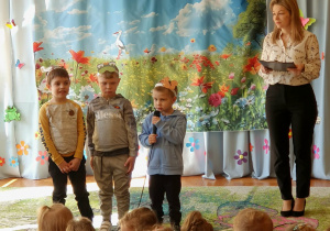 Na zdjęciu widać trzech chłopców stojących na scenie, którzy recytują wiersz przed zgromadzoną publicznością przedszkolną. Dzieci z zaangażowaniem prezentują swój występ, a pozostali uczestnicy z uwagą ich słuchają.