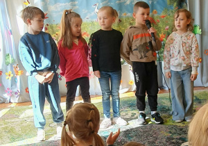 Na zdjęciu widać grupę dzieci stojących na scenie podczas przedszkolnego konkursu recytatorskiego o tematyce wiosennej. Dzieci ustawione są w rzędzie i wspólnie prezentują przygotowany utwór przed zgromadzoną publicznością. W tle widoczna jest dekoracja z napisem „Wiersz dla wiosny” oraz barwna plansza przedstawiająca wiosenną łąkę z kwiatami i ptakami. Scenę zdobią również motyle i inne elementy nawiązujące do budzącej się do życia przyrody.