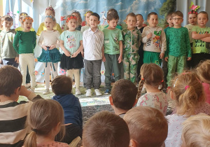 Na zdjęciu widać grupę dzieci stojących na scenie podczas przedszkolnego konkursu recytatorskiego o tematyce wiosennej. Dzieci ustawione są w rzędzie i wspólnie prezentują przygotowany utwór przed zgromadzoną publicznością. Ubrane są w kolorowe, wiosenne stroje – dominują odcienie zieleni oraz dodatki nawiązujące do przyrody, takie jak opaski z kwiatami. W tle widoczna jest dekoracja z napisem „Wiersz dla wiosny” oraz barwna plansza przedstawiająca wiosenną łąkę z kwiatami i ptakami. Scenę zdobią również motyle i inne elementy nawiązujące do budzącej się do życia przyrody. Na pierwszym planie siedzą dzieci – widownia, które z zainteresowaniem i skupieniem oglądają występ swoich kolegów. Cała scena ma radosny, uroczysty charakter, a atmosfera wydarzenia sprzyja wspólnemu przeżywaniu występów i rozwijaniu dziecięcych talentów.