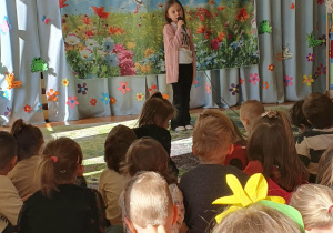 Na zdjęciu widać dziewczynkę stojącą na scenie, która recytuje wiersz, trzymając w ręku mikrofon. W tle znajduje się wiosenna dekoracja z napisem „Wiersz dla wiosny”, a przed sceną siedzą dzieci, które z uwagą słuchają występu.